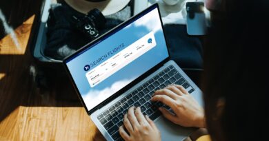 Young Asian woman shopping online for flight ticket on airline website with laptop at home, sitting next to suitcase with sun hat, camera, clothings, headphones, smartphone and passport. Getting ready for vacation. Travel and vacation concept